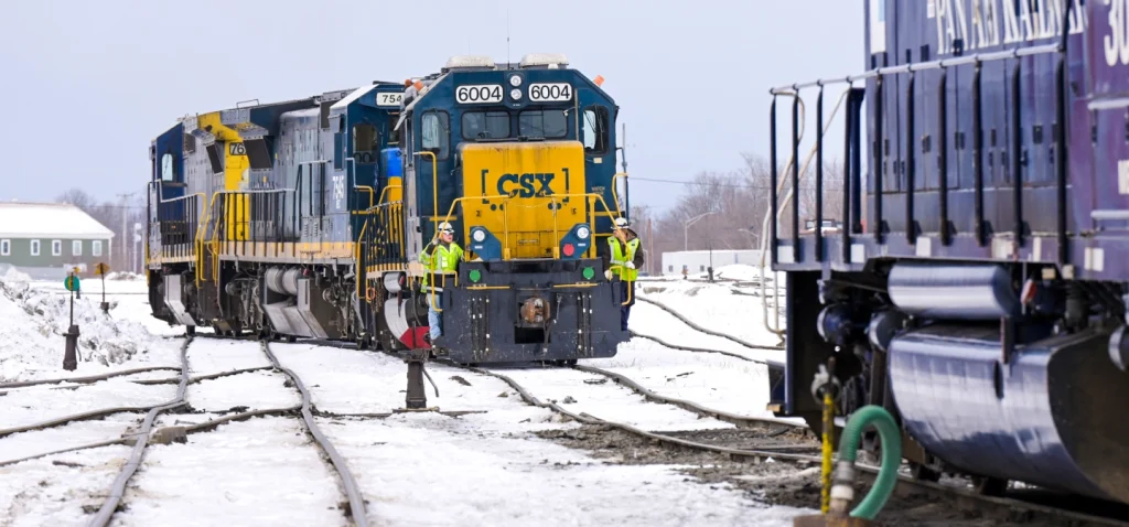 Two freight trains with workers standing on the front platforms are positioned on snowy railway tracks. The trains are painted in blue and yellow (CSX) and dark blue (Pan Am Railways). Snow covers the ground and tracks, and a green building is visible in the background.
